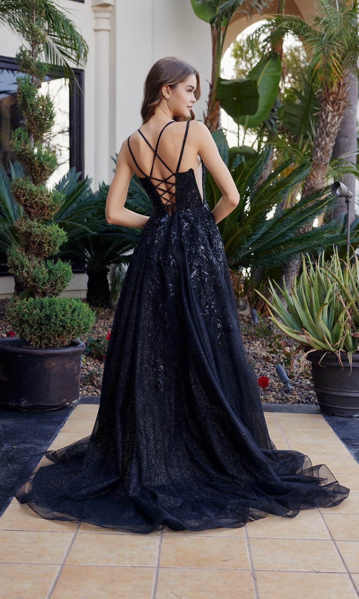 A woman stands outdoors among potted plants and palms, wearing the Nox Anabel H1885—a sleeveless long prom dress with a corset bodice and lace-up back.